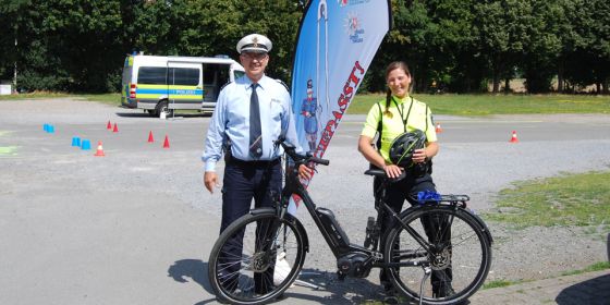 Im Hintergrund ein Streifenwagen. Ein Polizist sowie eine Polizistin stehen vor einem aufgebauten Pacour mit einem Fahrrad. 