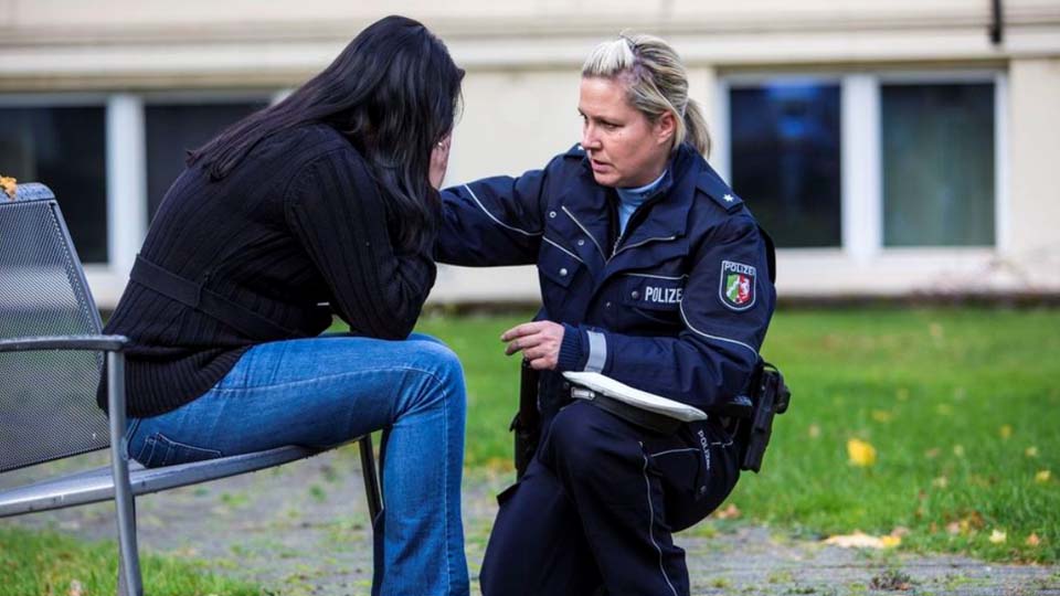 Eine Frau sitzt auf einem Stuhl und verdeckt ihr Gesicht. Eine Polizistin kniet vor ihr und spricht mit ihr. 