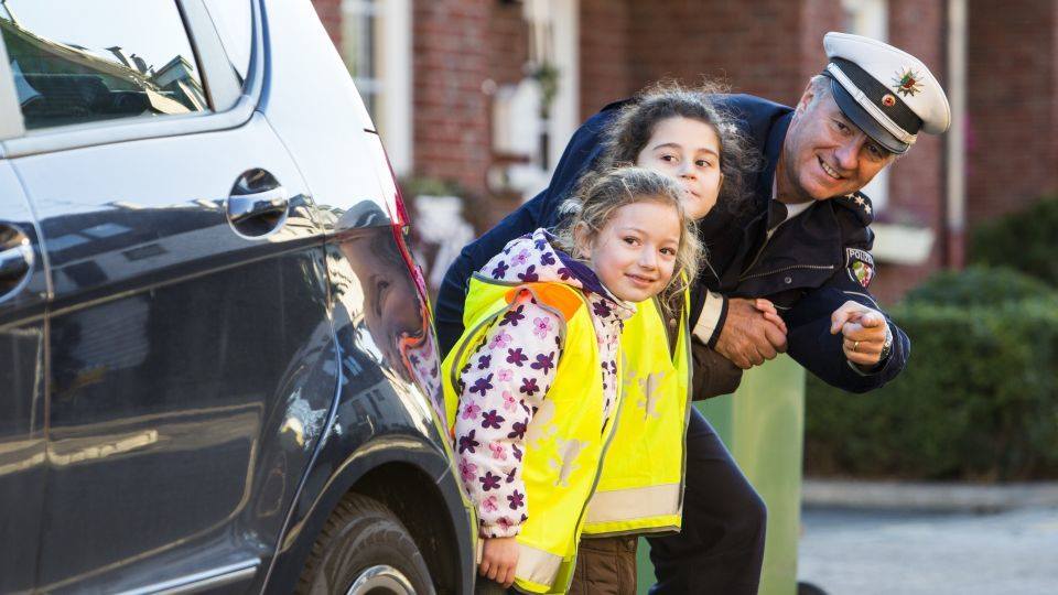 Zwei Kinder stehen neben einem Auto an der Straße. Ein Polizist steht daneben und zeigt auf etwas. 