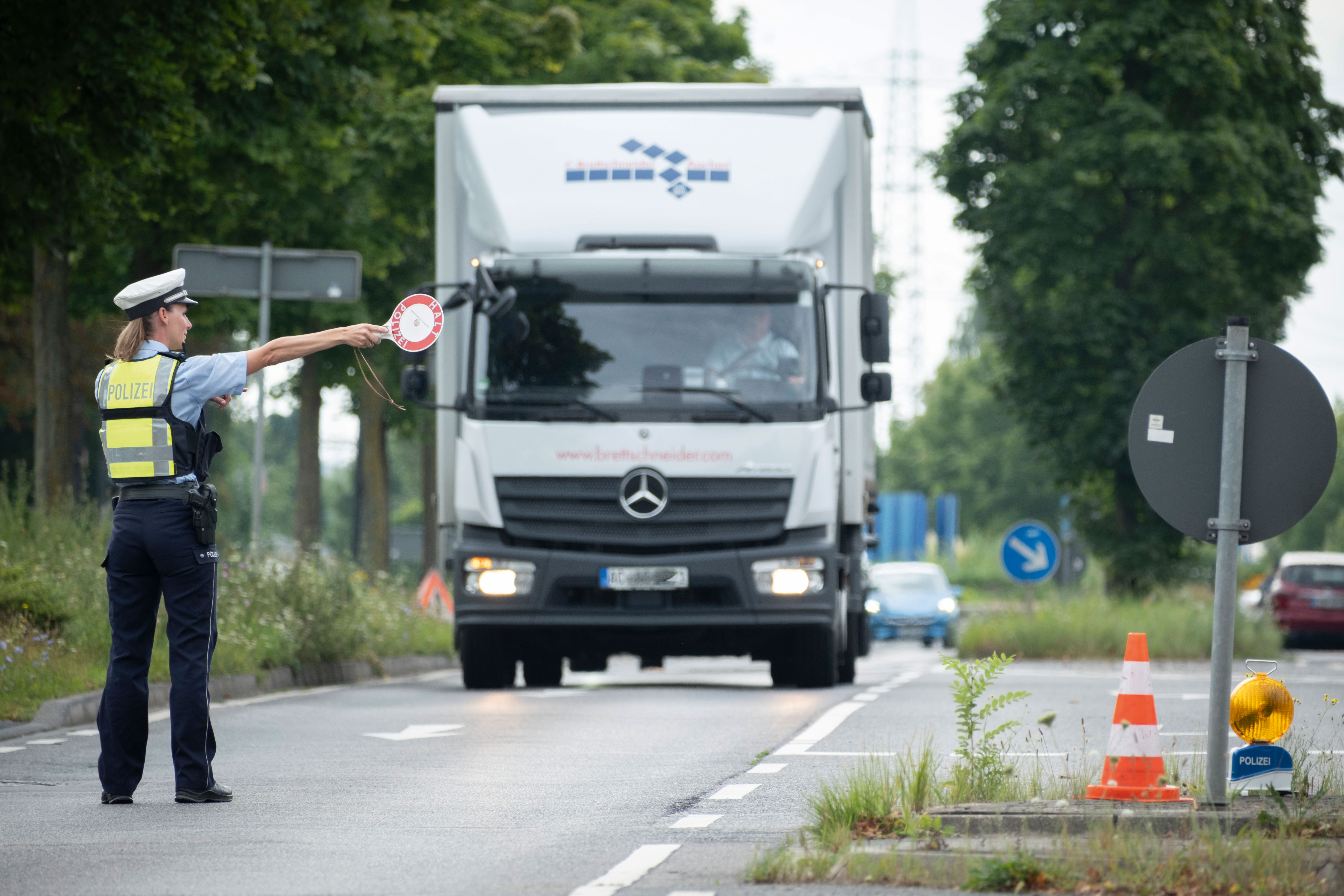 On the left, uniformed policewoman with a police cap on her head and a red and white stop trowel in her right hand. She is standing on the road. A white LKA with a blue logo above the driver's cab rolls towards her. Her arm with the stop trowel is pointing to the right towards a parking lot.