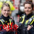 Im Vordergrund sind die Polizistinnen Paula Petzschke und Jennifer Krystkiewicz zu sehen. Sie tragen Uniform und haben die Hände an ihrer Weste eingehakt. Beide schauen in die Kamera. Im Hintergrund sind Rettungskräfte zu sehen.