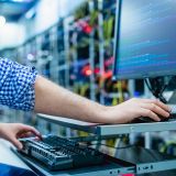 Im Hintergrund ist schemenhaft ein Serverraum zu sehen. Vorne sitzt ein Mann an einem Rechner. 