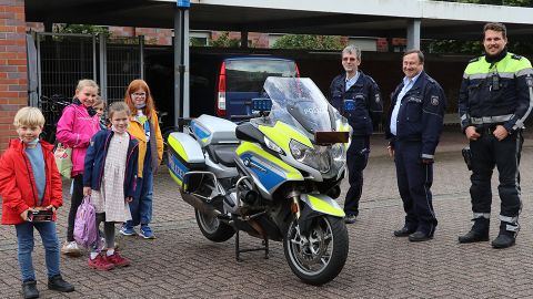 Fünf Kinder stehen neben einem Polizeimotorrad. Rechts daneben stehen drei Polizisten. 