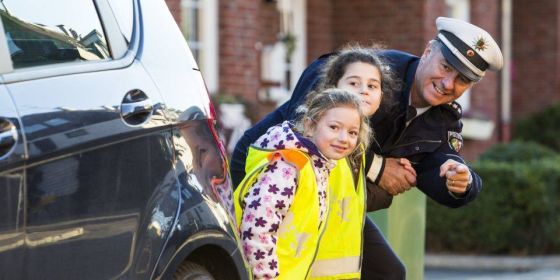 Zwei Kinder stehen neben einem Auto an der Straße. Ein Polizist steht daneben und zeigt auf etwas. 
